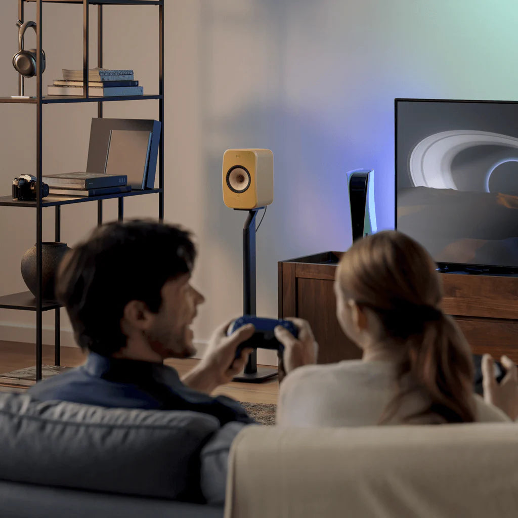 Two people sitting on a couch playing video games in a living room with a TV and speakers.